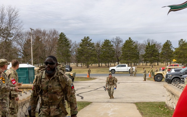 Military personnel in Army Combat Uniforms (ACU/OCP pattern) are participating in what appears to be a ruck march or physical fitness event at a military installation. A lead soldier carrying a rucksack and rifle is walking along a paved pathway while fellow soldiers line the sides, appearing to offer encouragement. A red and gold military unit guidon flag is visible on the right side of the frame.