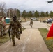 Military personnel in Army Combat Uniforms (ACU/OCP pattern) are participating in what appears to be a ruck march or physical fitness event at a military installation. A lead soldier carrying a rucksack and rifle is walking along a paved pathway while fellow soldiers line the sides, appearing to offer encouragement. A red and gold military unit guidon flag is visible on the right side of the frame.