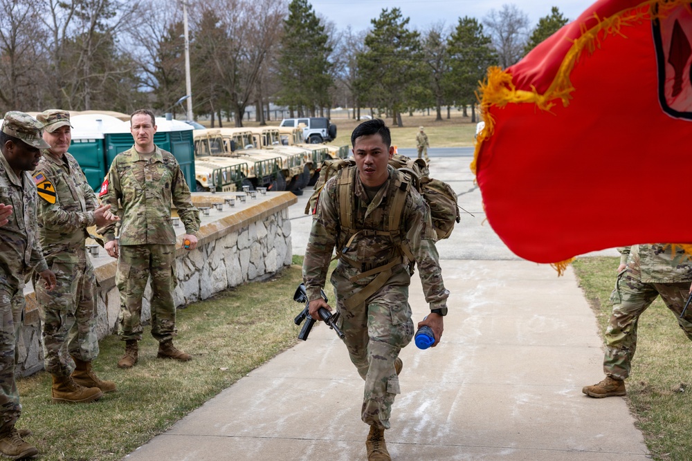 A U.S. Army soldier in full combat gear, carrying a rifle and water bottle with a rucksack, appears to be completing a ruck march or physical fitness event on a military installation. Fellow soldiers and officers line the walkway, applauding and observing the participant. A red military guidon flag with gold fringe is visible in the foreground right.