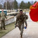 A U.S. Army soldier in full combat gear, carrying a rifle and water bottle with a rucksack, appears to be completing a ruck march or physical fitness event on a military installation. Fellow soldiers and officers line the walkway, applauding and observing the participant. A red military guidon flag with gold fringe is visible in the foreground right.