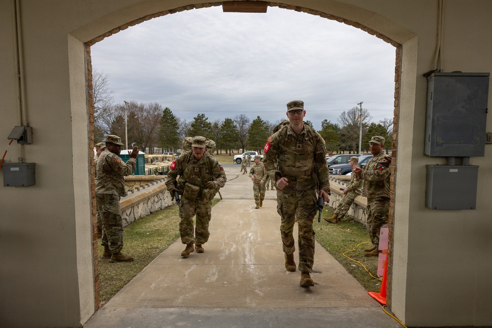 Military personnel in U.S. Army camouflage (OCP/Multicam) uniforms are walking through a brick archway at what appears to be a military installation. Several soldiers are wearing rucksacks and red arm bands, suggesting a training event or ruck march activity. The setting appears to be a well-maintained base facility with a parking area and tree-lined grounds visible in the background.