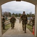 Military personnel in U.S. Army camouflage (OCP/Multicam) uniforms are walking through a brick archway at what appears to be a military installation. Several soldiers are wearing rucksacks and red arm bands, suggesting a training event or ruck march activity. The setting appears to be a well-maintained base facility with a parking area and tree-lined grounds visible in the background.