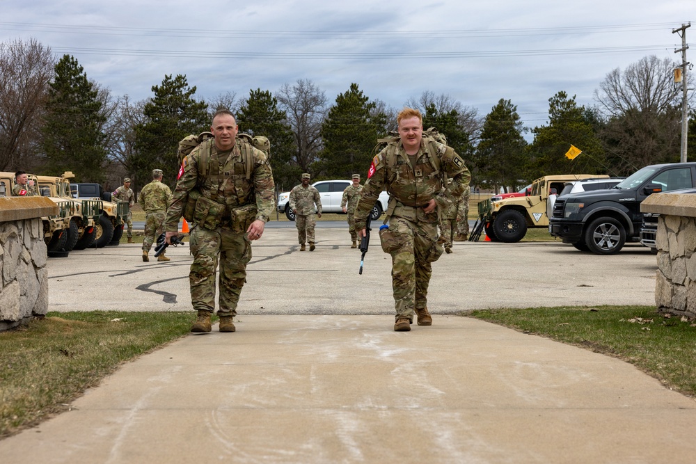 Two U.S. Army soldiers in OCP (Operational Camouflage Pattern) uniforms are walking toward the camera carrying large rucksacks, likely completing a ruck march event. Both soldiers wear red patches on their left shoulders and appear to be at a military installation with Humvees and civilian vehicles visible in the background. Additional soldiers and personnel are visible in the background along a paved pathway.