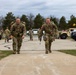 Two U.S. Army soldiers in OCP (Operational Camouflage Pattern) uniforms are walking toward the camera carrying large rucksacks, likely completing a ruck march event. Both soldiers wear red patches on their left shoulders and appear to be at a military installation with Humvees and civilian vehicles visible in the background. Additional soldiers and personnel are visible in the background along a paved pathway.