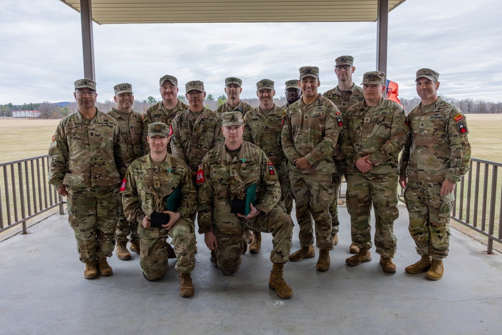 A group of approximately 13 military personnel in U.S. Army OCP (Operational Camouflage Pattern) uniforms pose for a formal group photograph under a covered outdoor pavilion. Two soldiers are kneeling in the front row, with one visibly wearing a medal and both holding what appear to be award cases or folders. The setting appears to be a military installation with open fields and buildings visible in the background.
