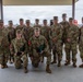 A group of approximately 13 military personnel in U.S. Army OCP (Operational Camouflage Pattern) uniforms pose for a formal group photograph under a covered outdoor pavilion. Two soldiers are kneeling in the front row, with one visibly wearing a medal and both holding what appear to be award cases or folders. The setting appears to be a military installation with open fields and buildings visible in the background.