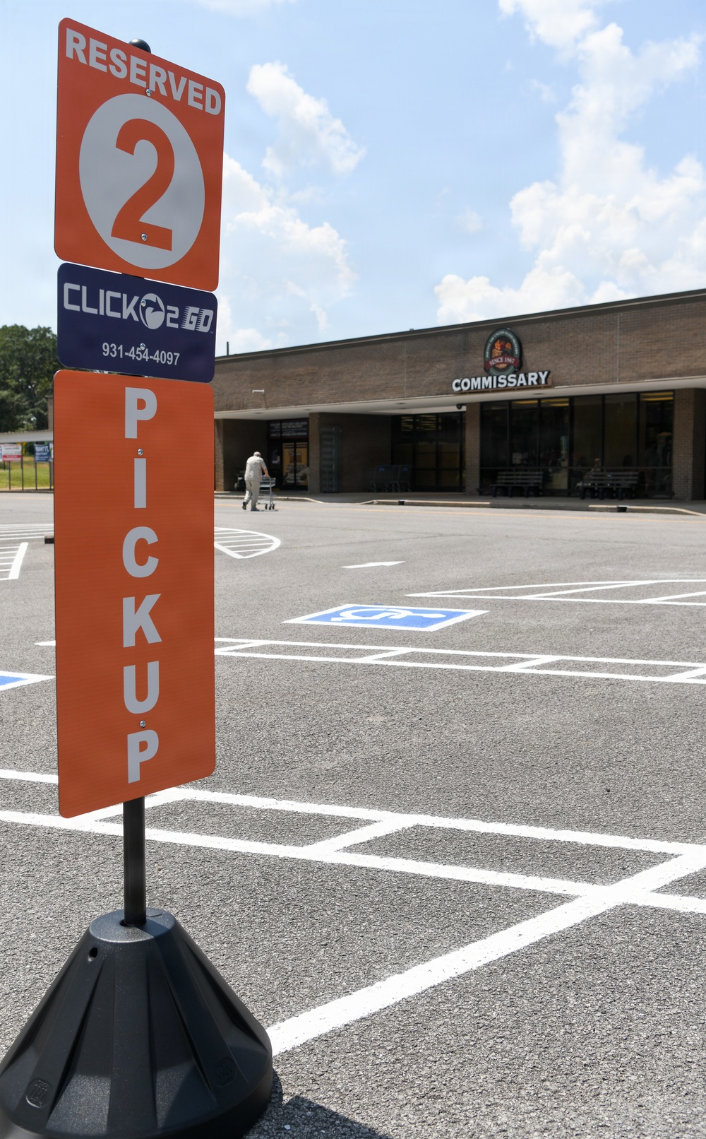 A parking lot sign for a 'Click2Go' curbside pickup service is prominently displayed in the foreground, marking Reserved Spot 2 at a military Commissary grocery store. The sign is orange and blue, mounted on a weighted black base, with a phone number (931-454-4097) visible. In the background, a person with a shopping cart is walking toward the store entrance.