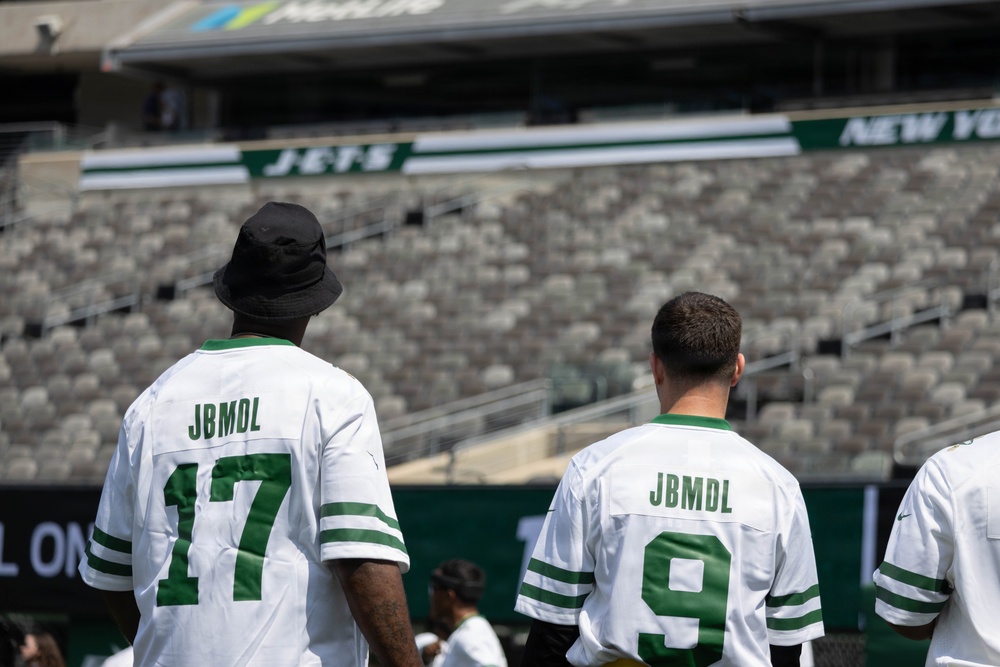 Two individuals are standing with their backs to the camera inside a New York Jets NFL stadium, wearing matching white Jets-style jerseys with 'JBMDL' printed on the back and numbers 17 and 9. The stadium seats are largely empty, suggesting this may be a pre-game, practice, or special event. The green and white Jets branding is clearly visible on the stadium signage in the background.