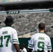 Two individuals are standing with their backs to the camera inside a New York Jets NFL stadium, wearing matching white Jets-style jerseys with 'JBMDL' printed on the back and numbers 17 and 9. The stadium seats are largely empty, suggesting this may be a pre-game, practice, or special event. The green and white Jets branding is clearly visible on the stadium signage in the background.