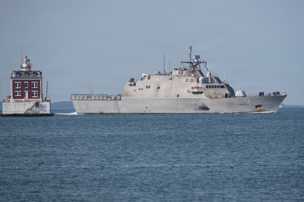 A U.S. Navy Littoral Combat Ship bearing hull number 13 is underway on open water, passing a red-brick lighthouse structure on the left. The vessel displays the characteristic angular, low-profile design of the Freedom-class LCS. Personnel are visible on the ship's deck, suggesting a transit or commissioning event.