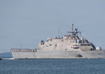 A U.S. Navy Littoral Combat Ship bearing hull number 13 is underway on open water, passing a red-brick lighthouse structure on the left. The vessel displays the characteristic angular, low-profile design of the Freedom-class LCS. Personnel are visible on the ship's deck, suggesting a transit or commissioning event.