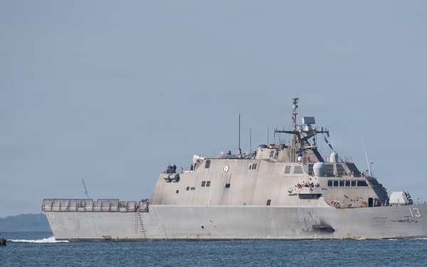 A U.S. Navy Littoral Combat Ship bearing hull number 13 is underway on open water, passing a red-brick lighthouse structure on the left. The vessel displays the characteristic angular, low-profile design of the Freedom-class LCS. Personnel are visible on the ship's deck, suggesting a transit or commissioning event.