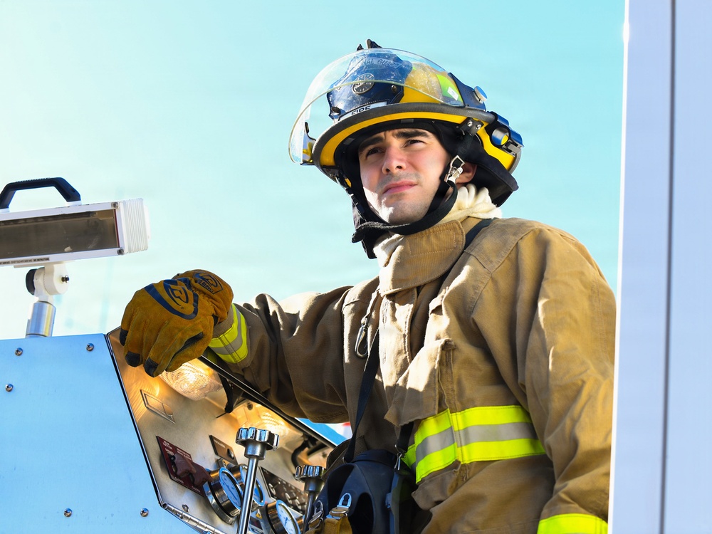 A young male firefighter is positioned on or near a fire apparatus, operating equipment controls. He is wearing full structural firefighting gear including a yellow and black helmet with a face shield, tan turnout coat with yellow reflective striping, and leather work gloves. The image appears to be taken during active operations or a training exercise.