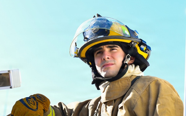 A young male firefighter is positioned on or near a fire apparatus, operating equipment controls. He is wearing full structural firefighting gear including a yellow and black helmet with a face shield, tan turnout coat with yellow reflective striping, and leather work gloves. The image appears to be taken during active operations or a training exercise.
