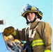 A young male firefighter is positioned on or near a fire apparatus, operating equipment controls. He is wearing full structural firefighting gear including a yellow and black helmet with a face shield, tan turnout coat with yellow reflective striping, and leather work gloves. The image appears to be taken during active operations or a training exercise.