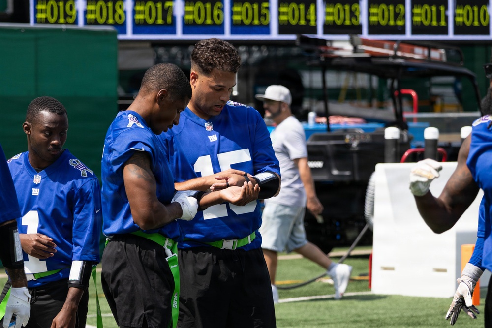 Several players wearing New York Giants blue NFL jerseys are gathered on a sideline or practice field. The player wearing jersey number 15 appears to be reviewing a wristband playbook or electronic device with a teammate. Green flag football belts are visible, suggesting this may be a flag football or non-contact practice session.