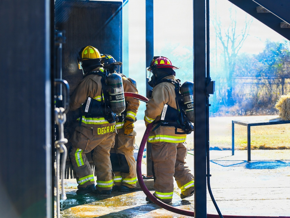 Three firefighters in full turnout gear and SCBA (Self-Contained Breathing Apparatus) are entering or positioned at the threshold of a structure, holding a charged hose line. The scene appears to be a live fire training exercise, evidenced by the controlled environment, smoke visible in the background, and the structured setting. One firefighter's gear is labeled 'DEGRAF' on the back of the pants.