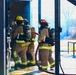 Three firefighters in full turnout gear and SCBA (Self-Contained Breathing Apparatus) are entering or positioned at the threshold of a structure, holding a charged hose line. The scene appears to be a live fire training exercise, evidenced by the controlled environment, smoke visible in the background, and the structured setting. One firefighter's gear is labeled 'DEGRAF' on the back of the pants.