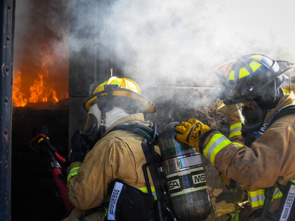 Two firefighters in full personal protective equipment are approaching an active fire through a smoke-filled doorway or opening. Both are wearing SCBA (Self-Contained Breathing Apparatus) units branded 'MSA' and full turnout gear with high-visibility reflective striping. The scene appears to be a live fire training exercise or active structural fire response.