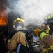 Two firefighters in full personal protective equipment are approaching an active fire through a smoke-filled doorway or opening. Both are wearing SCBA (Self-Contained Breathing Apparatus) units branded 'MSA' and full turnout gear with high-visibility reflective striping. The scene appears to be a live fire training exercise or active structural fire response.