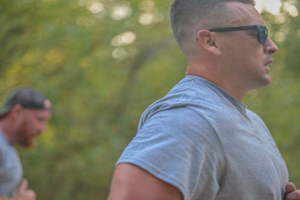 Two men are outdoors in a wooded or tree-lined setting, appearing to be engaged in physical activity such as running or walking. The foreground subject is a muscular man wearing a gray t-shirt and dark sunglasses with a short military-style haircut. A second man wearing a baseball cap is visible but out of focus in the background.