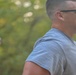 Two men are outdoors in a wooded or tree-lined setting, appearing to be engaged in physical activity such as running or walking. The foreground subject is a muscular man wearing a gray t-shirt and dark sunglasses with a short military-style haircut. A second man wearing a baseball cap is visible but out of focus in the background.