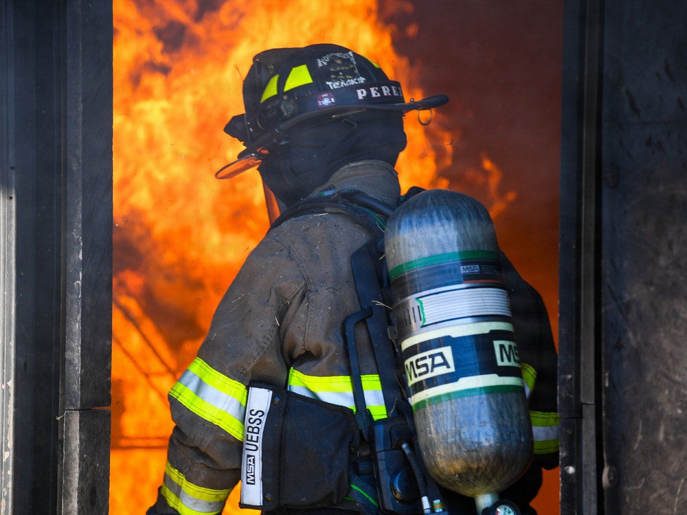 A firefighter wearing full personal protective equipment stands at the threshold of a doorway engulfed in intense orange flames. The firefighter is equipped with an MSA self-contained breathing apparatus (SCBA) and turnout gear bearing the name 'PEREZ' on the helmet. The image captures a high-risk moment of active firefighting operations.