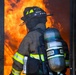A firefighter wearing full personal protective equipment stands at the threshold of a doorway engulfed in intense orange flames. The firefighter is equipped with an MSA self-contained breathing apparatus (SCBA) and turnout gear bearing the name 'PEREZ' on the helmet. The image captures a high-risk moment of active firefighting operations.
