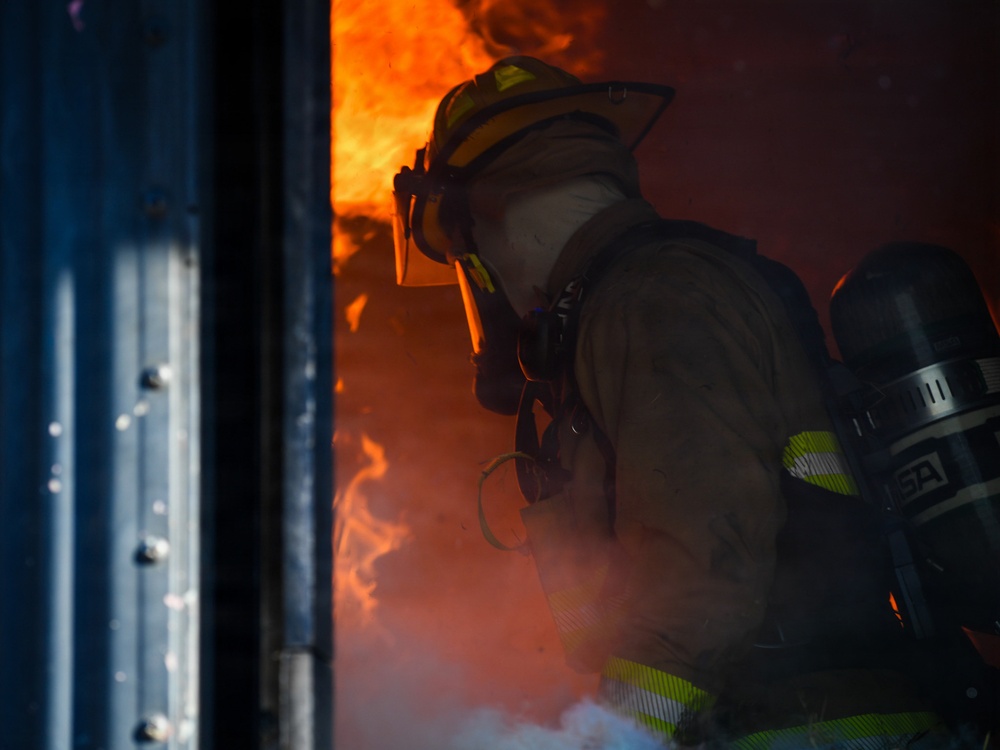 A firefighter in full protective gear is positioned at the threshold of a doorway engulfed in intense flames and smoke. The firefighter is crouched low, wearing a yellow helmet, turnout gear, and an MSA self-contained breathing apparatus (SCBA). The scene depicts active firefighting operations with visible fire and heavy smoke conditions.