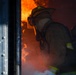 A firefighter in full protective gear is positioned at the threshold of a doorway engulfed in intense flames and smoke. The firefighter is crouched low, wearing a yellow helmet, turnout gear, and an MSA self-contained breathing apparatus (SCBA). The scene depicts active firefighting operations with visible fire and heavy smoke conditions.