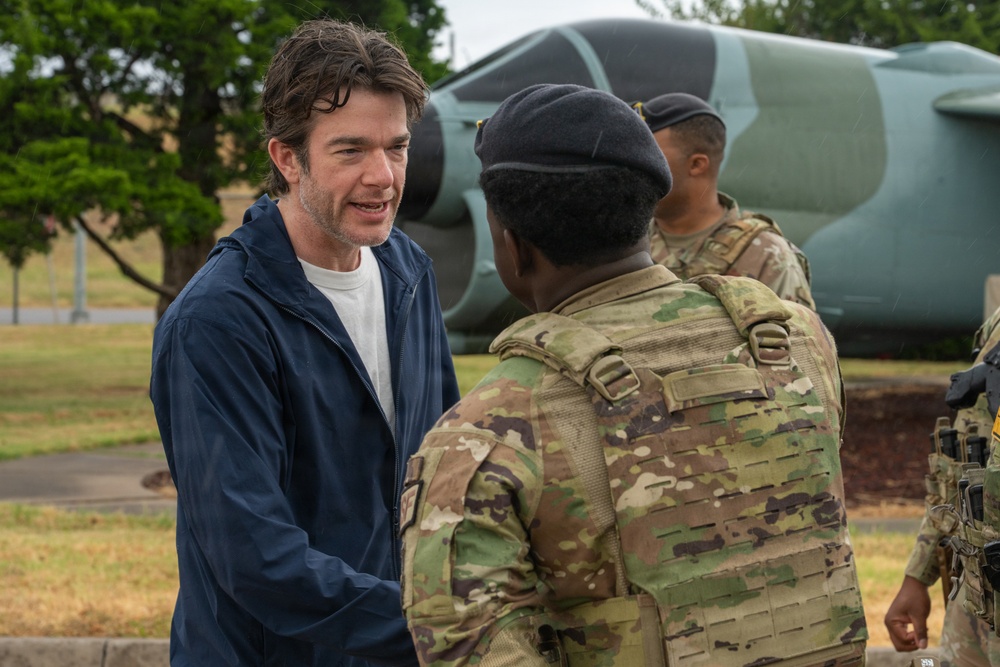 A civilian man in a navy blue jacket is shaking hands or greeting a military soldier in full camouflage tactical gear and a black beret. At least two additional soldiers in matching OCP (Operational Camouflage Pattern) uniforms are visible in the background. The setting appears to be a military installation, with what looks like a static display aircraft visible in the background.