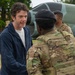 A civilian man in a navy blue jacket is shaking hands or greeting a military soldier in full camouflage tactical gear and a black beret. At least two additional soldiers in matching OCP (Operational Camouflage Pattern) uniforms are visible in the background. The setting appears to be a military installation, with what looks like a static display aircraft visible in the background.