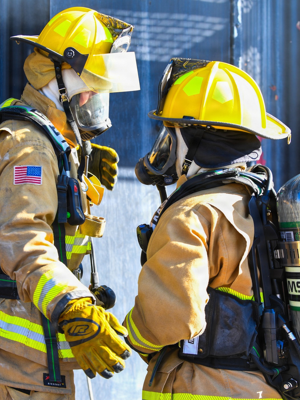 Two firefighters in full personal protective equipment face each other, appearing to communicate or coordinate during an operation. Both wear yellow helmets, turnout gear with high-visibility reflective striping, SCBA (Self-Contained Breathing Apparatus) units, and full-face respirator masks. An American flag patch is visible on the left firefighter's sleeve, and SCBA equipment is branded 'MSA' on the right firefighter's tank.