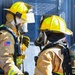 Two firefighters in full personal protective equipment face each other, appearing to communicate or coordinate during an operation. Both wear yellow helmets, turnout gear with high-visibility reflective striping, SCBA (Self-Contained Breathing Apparatus) units, and full-face respirator masks. An American flag patch is visible on the left firefighter's sleeve, and SCBA equipment is branded 'MSA' on the right firefighter's tank.