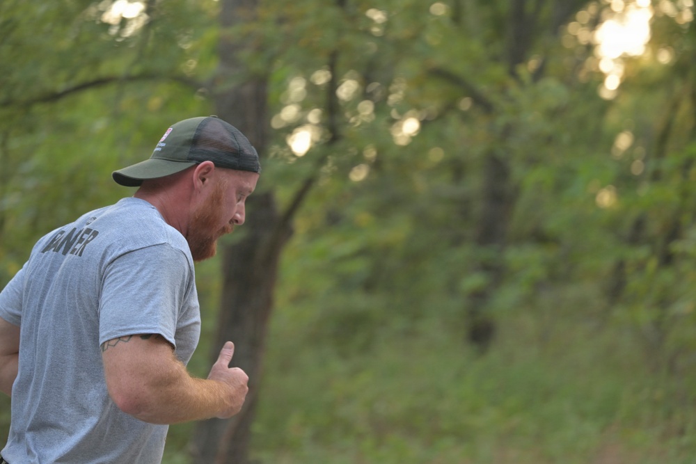 A man with a red beard is running or jogging outdoors in a wooded or park-like setting. He is wearing a gray t-shirt with partial text visible on the back reading 'ANER' and a green trucker-style baseball cap. The photo is taken from a side/rear angle during what appears to be early morning or late afternoon based on the warm, diffused lighting.