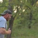 A man with a red beard is running or jogging outdoors in a wooded or park-like setting. He is wearing a gray t-shirt with partial text visible on the back reading 'ANER' and a green trucker-style baseball cap. The photo is taken from a side/rear angle during what appears to be early morning or late afternoon based on the warm, diffused lighting.