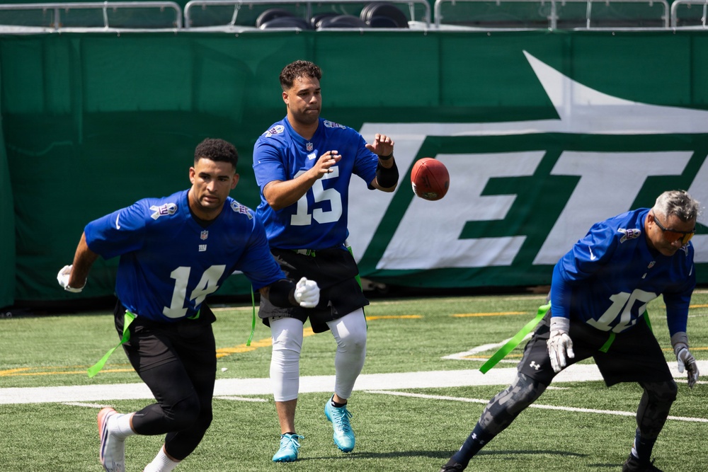 Three players in blue NFL jerseys are participating in a flag football game on a turf field. The central player wearing number 15 is in the act of throwing or releasing a football, while players numbered 14 and 10 are running nearby. The game appears to be taking place at MetLife Stadium, home of the New York Jets, as indicated by the large 'JETS' banner in the background.
