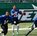 Three players in blue NFL jerseys are participating in a flag football game on a turf field. The central player wearing number 15 is in the act of throwing or releasing a football, while players numbered 14 and 10 are running nearby. The game appears to be taking place at MetLife Stadium, home of the New York Jets, as indicated by the large 'JETS' banner in the background.