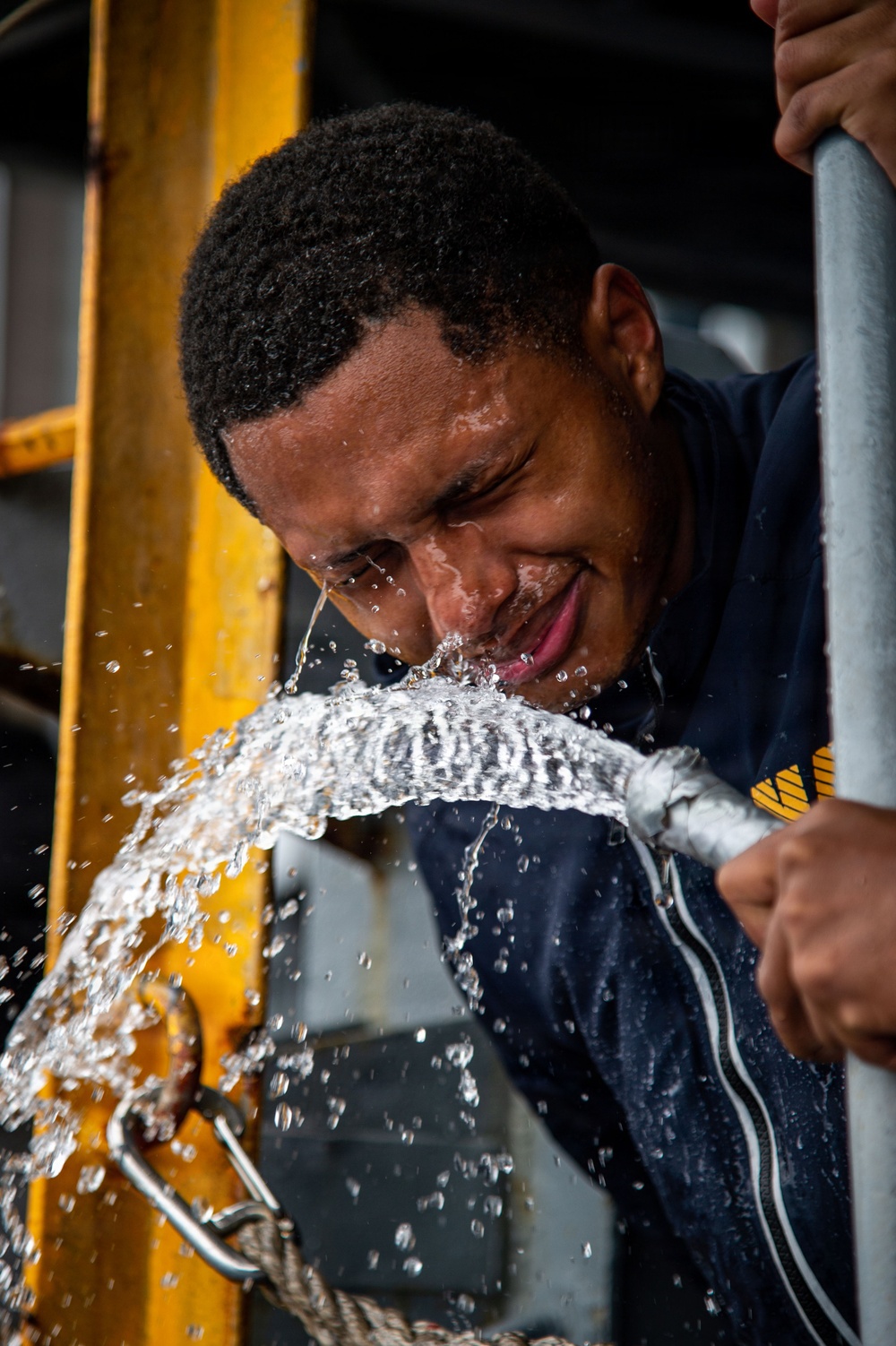 A young man in a dark navy blue shirt is leaning over a pressurized hose or pipe from which water is forcefully spraying, splashing across his face and chest. He appears to be smiling or grimacing as the water hits him. The setting appears to be an industrial or maritime environment with yellow structural framing visible in the background.