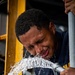 A young man in a dark navy blue shirt is leaning over a pressurized hose or pipe from which water is forcefully spraying, splashing across his face and chest. He appears to be smiling or grimacing as the water hits him. The setting appears to be an industrial or maritime environment with yellow structural framing visible in the background.