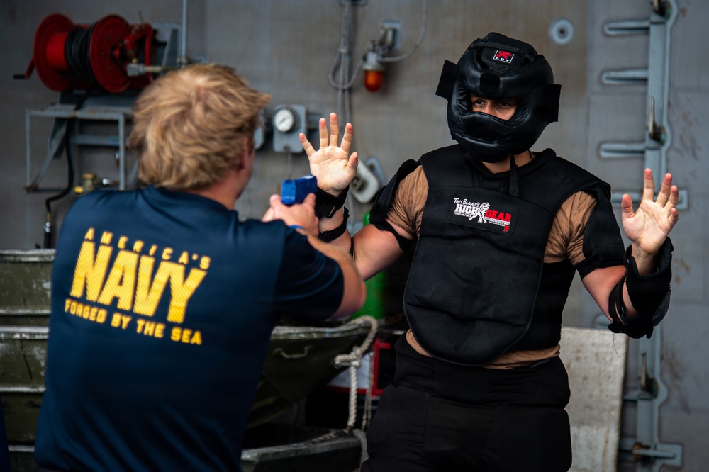 Two individuals are engaged in a training exercise aboard what appears to be a U.S. Navy vessel. One person wearing an 'America's Navy - Forged by the Sea' shirt is pointing a blue training device (likely a training taser or pepper spray) at a second person wearing full protective gear including a helmet, chest protector, and elbow pads. The person in protective gear has both hands raised in a surrender or defensive posture.