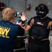 Two individuals are engaged in a training exercise aboard what appears to be a U.S. Navy vessel. One person wearing an 'America's Navy - Forged by the Sea' shirt is pointing a blue training device (likely a training taser or pepper spray) at a second person wearing full protective gear including a helmet, chest protector, and elbow pads. The person in protective gear has both hands raised in a surrender or defensive posture.
