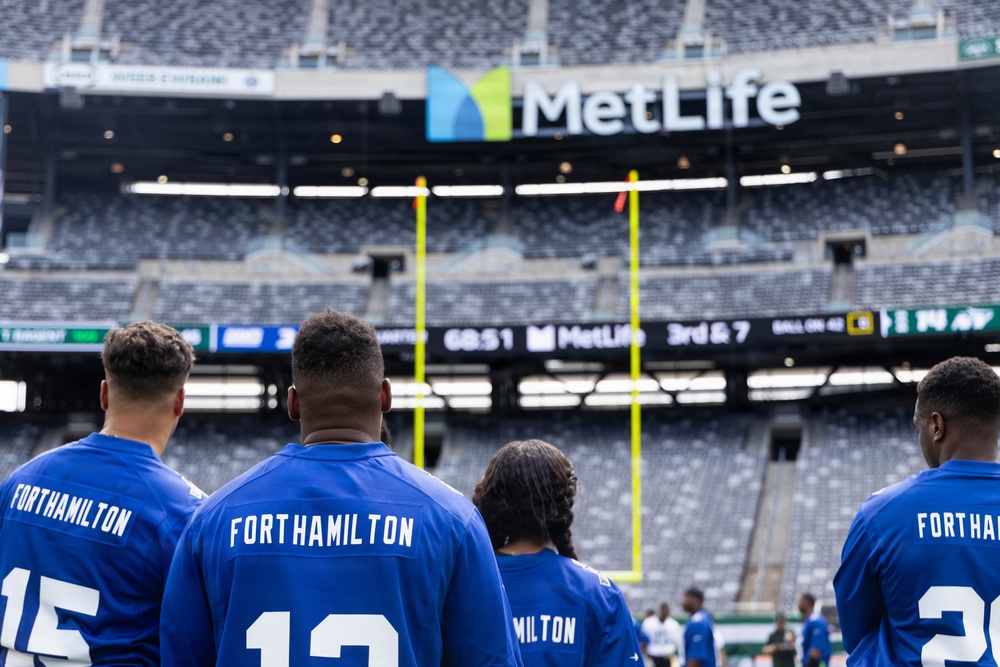 Four individuals wearing blue jerseys with 'FORTHAMILTON' printed on the back are standing on the field at MetLife Stadium, facing the field. The stadium appears largely empty, suggesting this is a pre-game or special event visit. The scoreboard displays '3rd &amp; 7' and a game clock reading '68:51'.