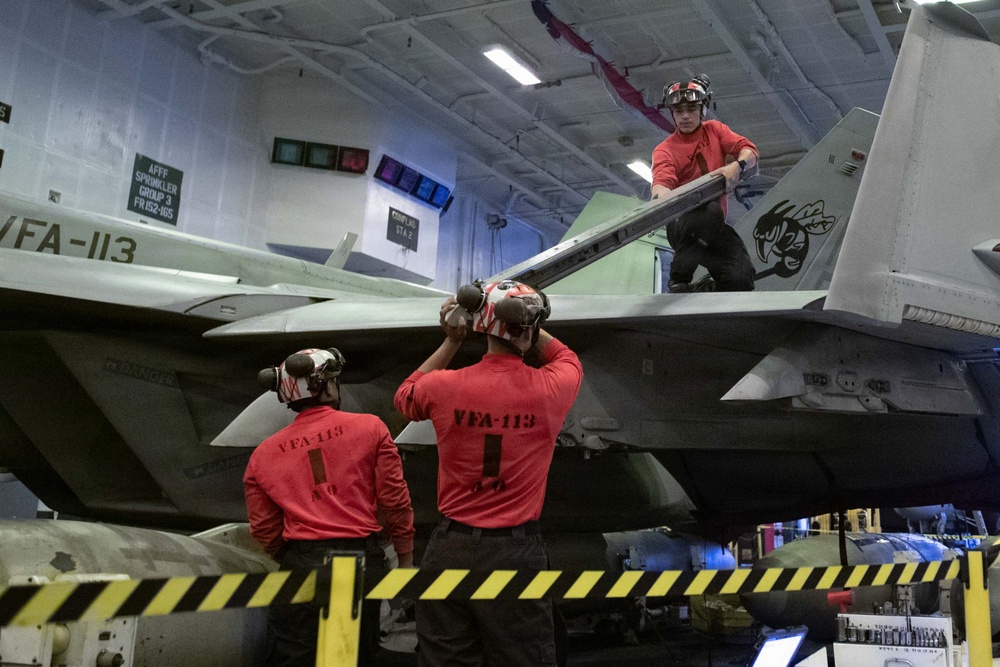 Three personnel in red uniforms marked 'VFA-113' are performing maintenance on a military jet aircraft, likely an F/A-18 Hornet or Super Hornet, inside an aircraft carrier hangar bay. Two crew members stand below the aircraft while a third is positioned on top of the wing. The hangar bay environment includes safety markings, signage, and equipment consistent with a U.S. Navy carrier air wing maintenance operation.