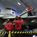 Three personnel in red uniforms marked 'VFA-113' are performing maintenance on a military jet aircraft, likely an F/A-18 Hornet or Super Hornet, inside an aircraft carrier hangar bay. Two crew members stand below the aircraft while a third is positioned on top of the wing. The hangar bay environment includes safety markings, signage, and equipment consistent with a U.S. Navy carrier air wing maintenance operation.