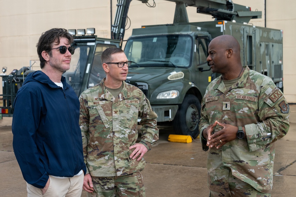 Three individuals are engaged in conversation on what appears to be a military installation or flight line. Two are wearing US Air Force OCP (Operational Camouflage Pattern) uniforms, while the third is a civilian in a navy blue hoodie. A military utility vehicle is visible in the background.