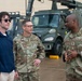 Three individuals are engaged in conversation on what appears to be a military installation or flight line. Two are wearing US Air Force OCP (Operational Camouflage Pattern) uniforms, while the third is a civilian in a navy blue hoodie. A military utility vehicle is visible in the background.