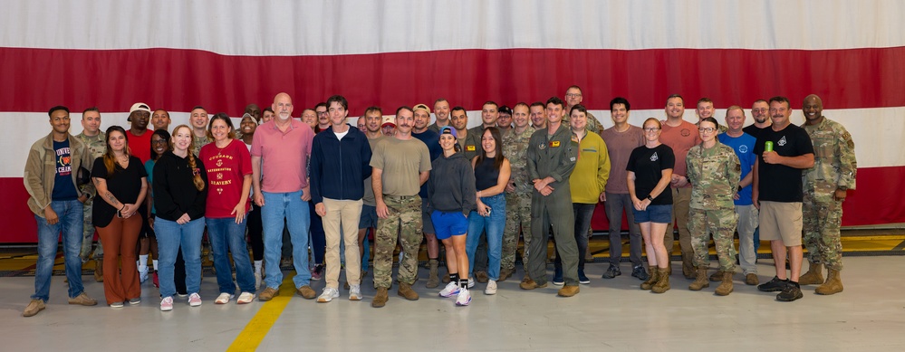A large group of approximately 35-40 individuals poses for a group photo inside what appears to be a military hangar. The group consists of a mix of military personnel in various uniforms (camouflage/OCP, flight suits) and civilians in casual attire. A large American flag serves as the backdrop behind the group.