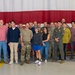 A large group of approximately 35-40 individuals poses for a group photo inside what appears to be a military hangar. The group consists of a mix of military personnel in various uniforms (camouflage/OCP, flight suits) and civilians in casual attire. A large American flag serves as the backdrop behind the group.