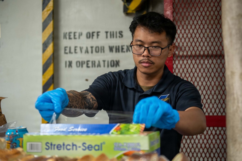 A young man wearing blue nitrile gloves and a dark polo shirt with a visible arm tattoo is handling food service materials near a 'Stretch-Seal' food wrap box. He is working in what appears to be an industrial or military vessel environment, indicated by the safety signage and metal grating in the background. The setting suggests a food service or galley operation.